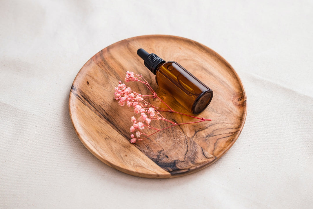 A dark brown dropper bottle next to a pinkish-red twig of flowers. They're sitting on a wooden plate with a marble, white background.