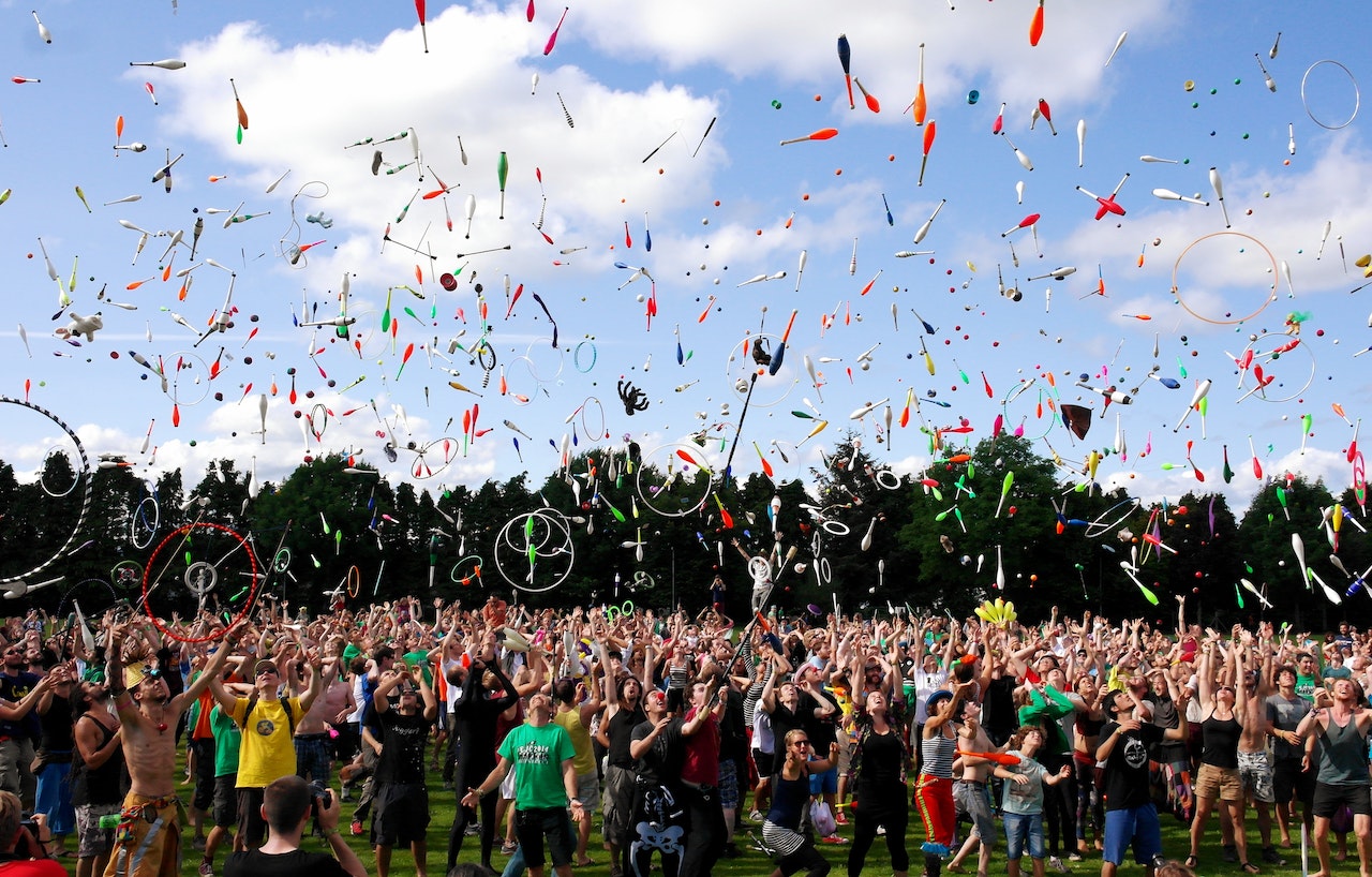 A festival crowd where everyone is throwing hula hoops and plastic, colorful bowling pins in the air.