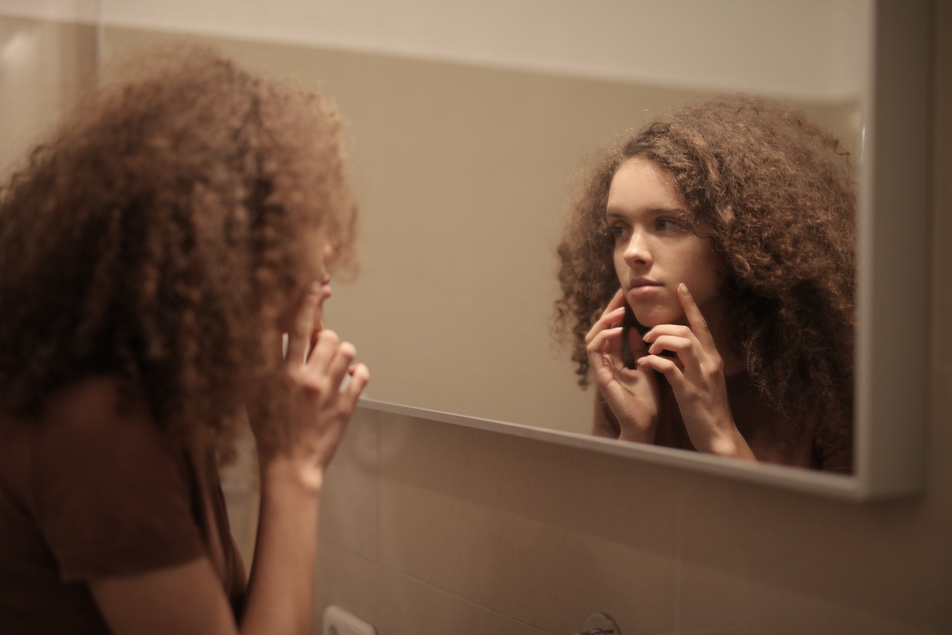 A teen girl with curly hair looks at her skin in a mirror. She looks unhappy.